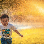 joyous boy running through a sprinkler with yellow summer light